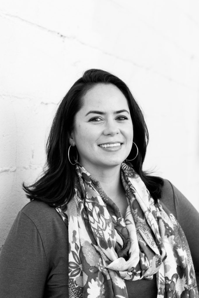 Black and white photo of the author, a smiling woman against a brick wall painted white, she is wearing a dark shirt and a white scarf with flowers on it.