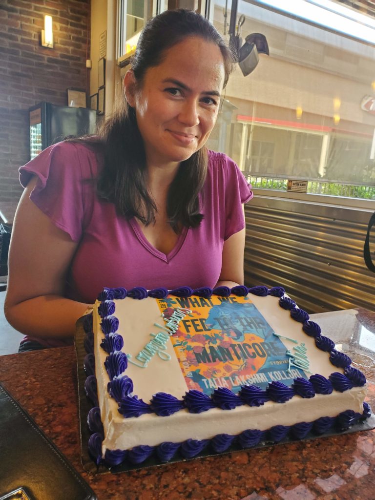 Talia sits at a table wearing a pink shirt. in front of her is a cake with her book cover printed on it. The cake says "Congratulations Talia"