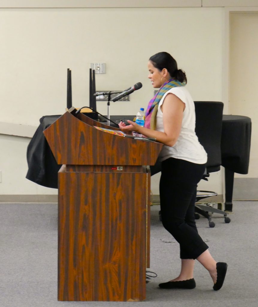 Talia stands at a podium in black pants, a white shirt and a multicolored scarf. She is reading from her book.